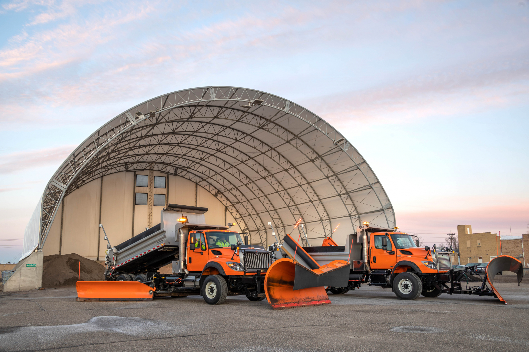 Snowplows sit in front of a stockpile of sand, salt, and other winter road necessities.