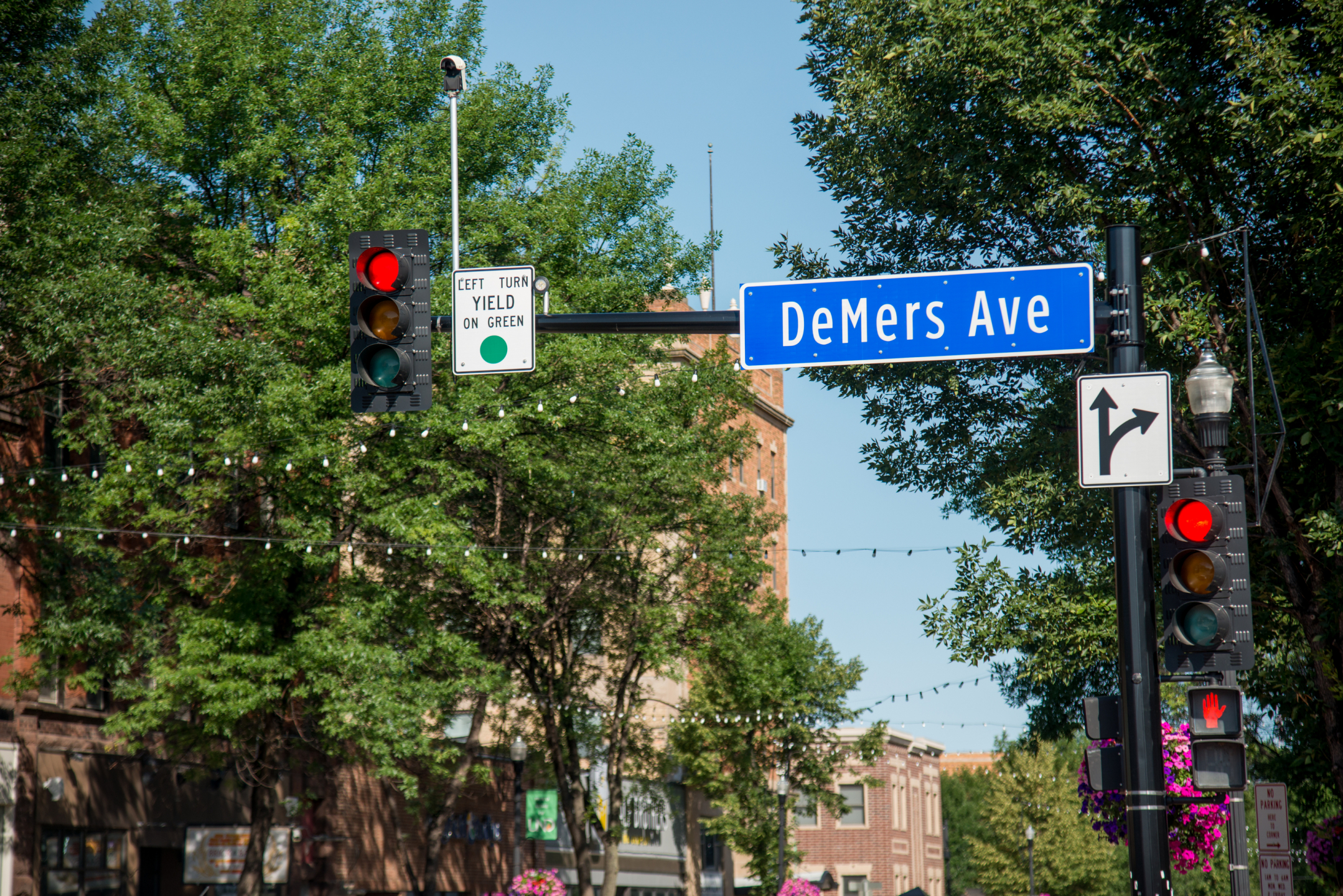 Image of DeMers Avenue sign on traffic light poles with trees and buildings in the background