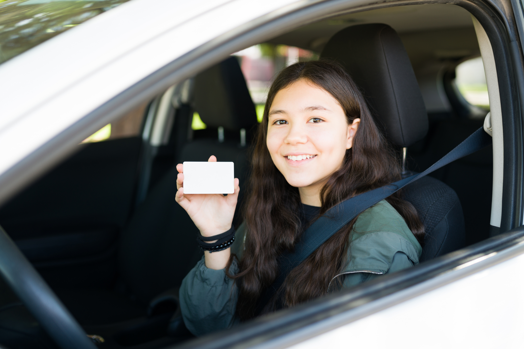 Smiling teenager looks out the window of a car while holding up a new driver license