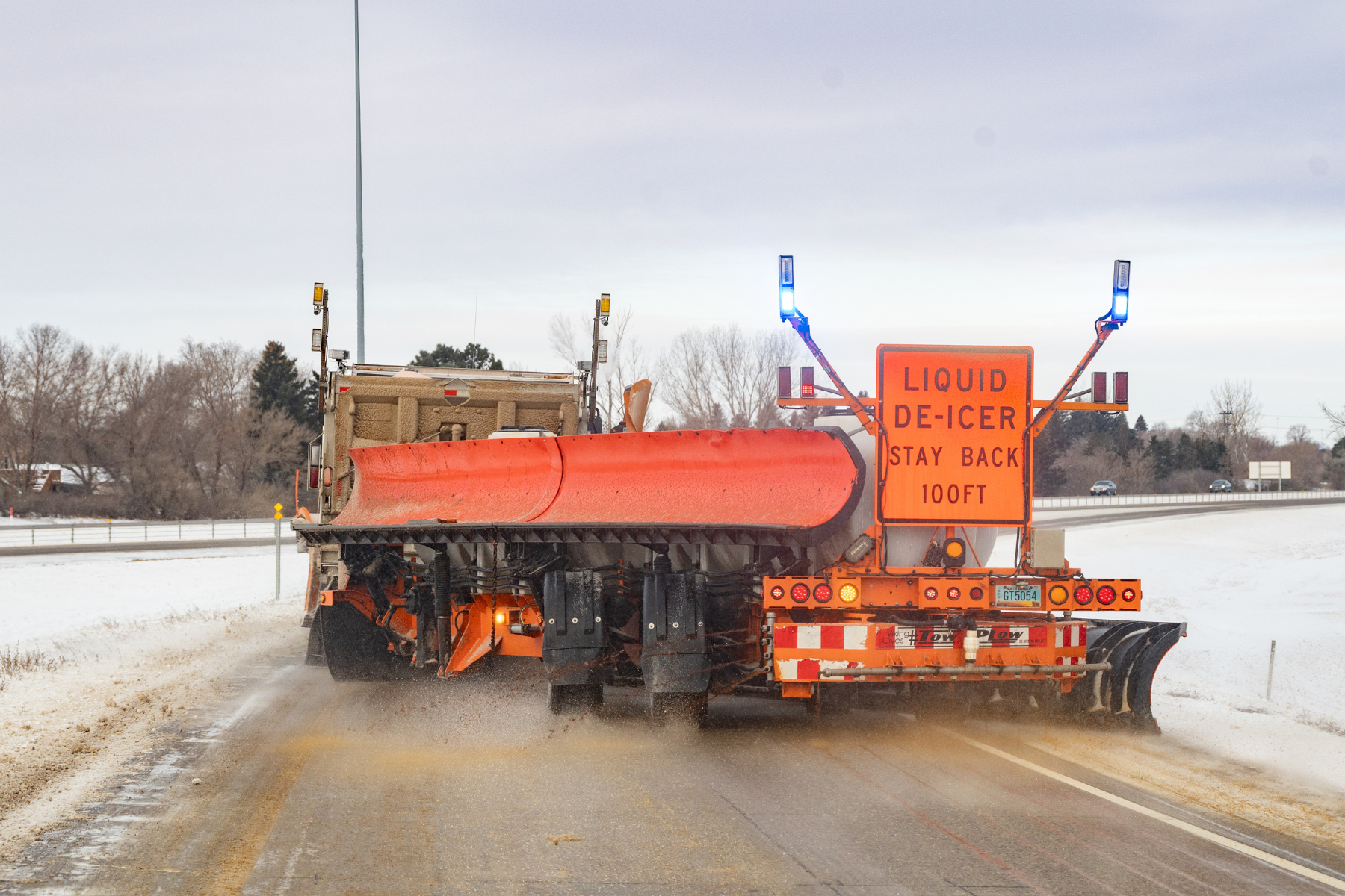 An NDDOT truck tows an extended plow with a brine tank and the sign on the back says Liquid De-Icer Stay Back 100 FT