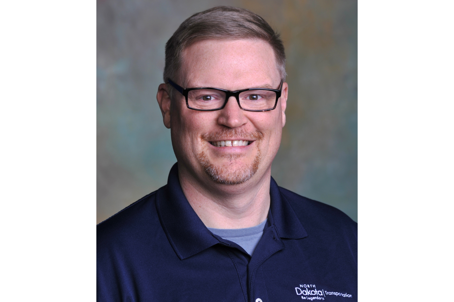 A headshot of Dan Hillerson. He has brown hair, rectangle glasses with dark frames, and a blue shirt.