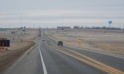 View entering US 85 to Belfield, ND, showing a 55 mph speed limit sign in the foreground, a vehicle on the highway, and a gas station with a water tower visible in the distance, surrounded by dry grass.