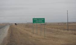 A portion of the US 85 roadway on the left with a 65 MPH speed sign and a Vision Zero road sign. Sky is gray with dry grass. 