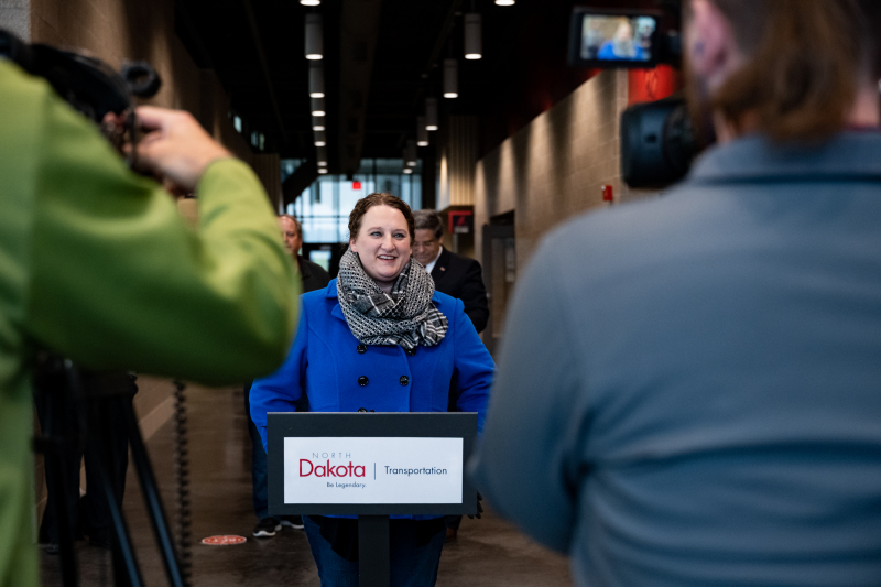 Women in blue coat and scarf stands at a podium with a NDDOT logo talking to reporters.